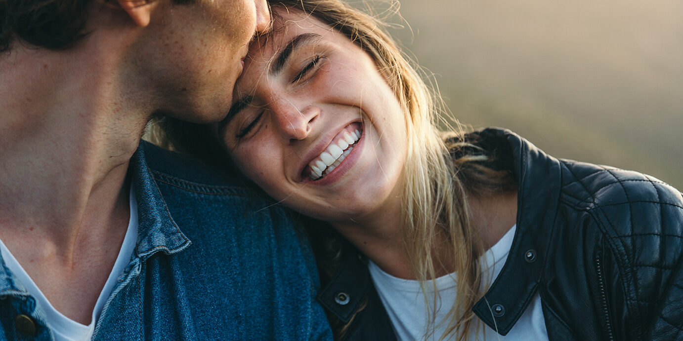 Close-up of romantic young boyfriend kissing smiling beautiful girlfriend on forehead while enjoying weekend at sunset