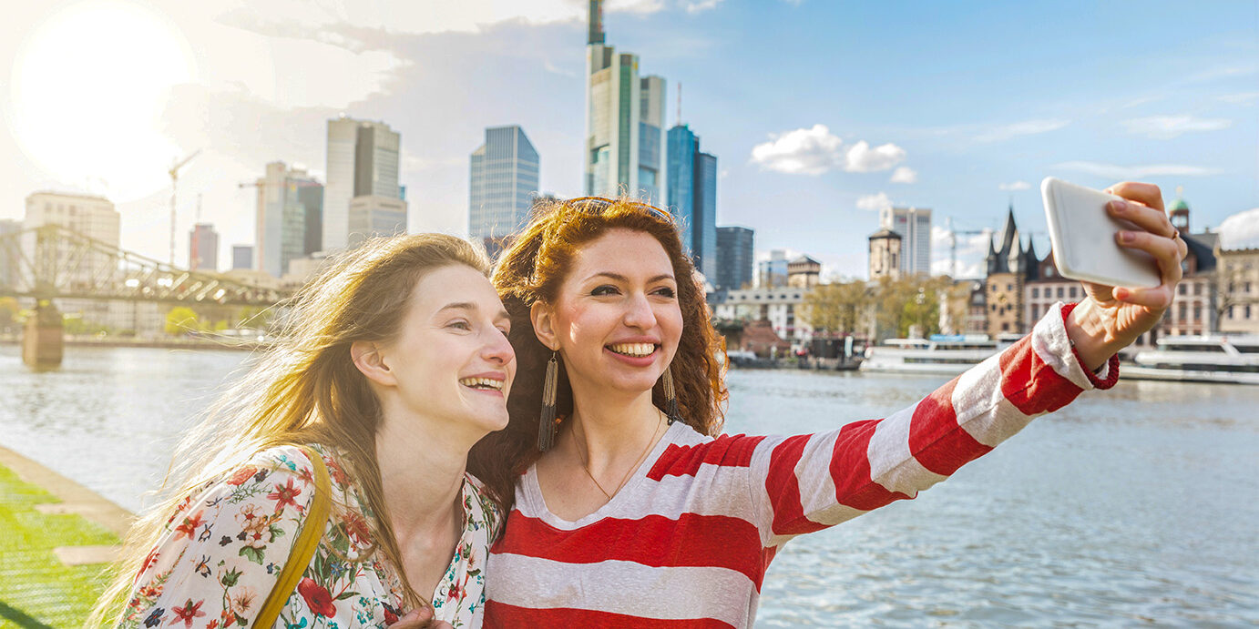 Two beautiful young women taking a selfie with Frankfurt skyline on background on a spring day. They are smiling and looking at the smart phone. Friendship and travel concepts.