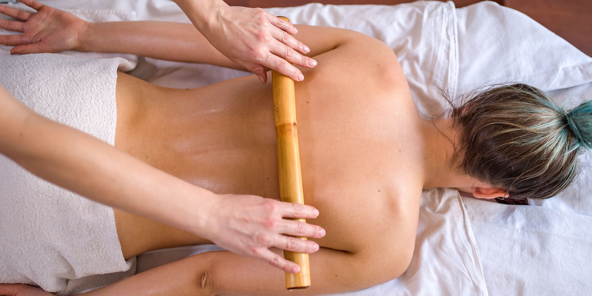 Woman is getting massage with bamboo stick in spa Young woman with green strands of hair is laying on front at massage table and getting bamboo stick massage by massage therapist.