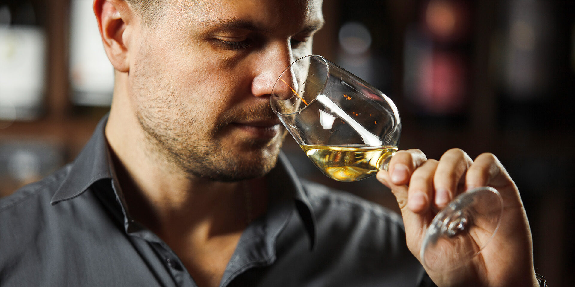 Bokal of white wine on background, male sommelier appreciating drink Sommelier smelling flavor of white wine in bokal on background of shelves with bottles in cellar. Male appreciating color, quality and sediments of drink. Professional degustation expert in winemaking.