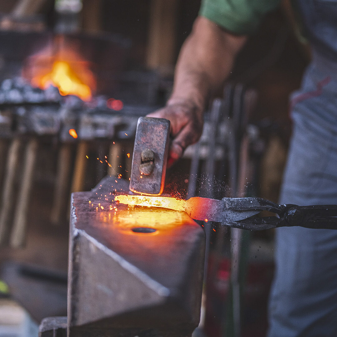 An unrecognizable blacksmith forges hot metal with a hammer on an anvile.