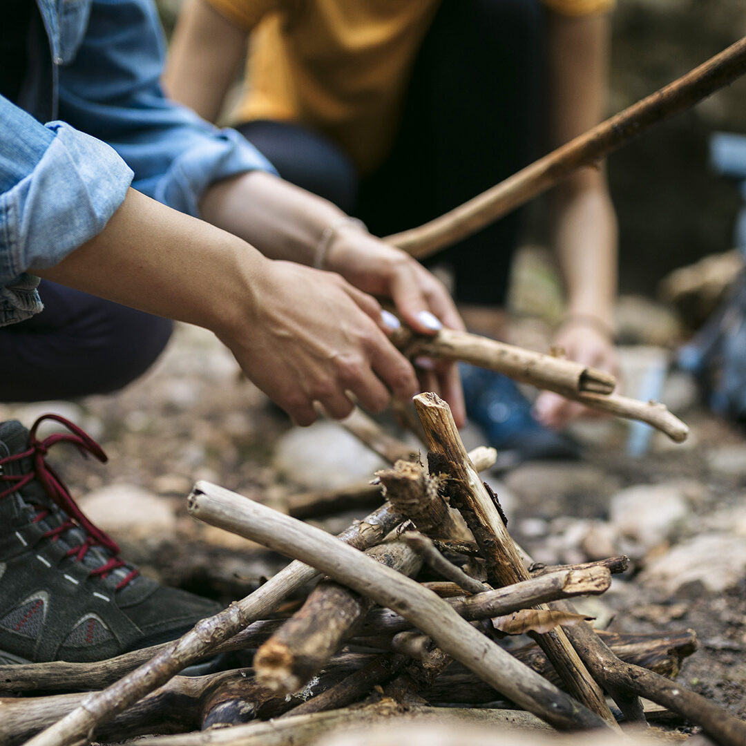 Close-up of two female hikers arranging sticks for campfire together for cooking food. Cropped shot of women hikers preparing campfire on forest trail.