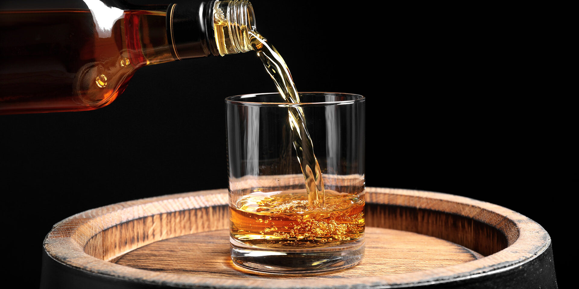 Pouring whiskey from bottle into glass on wooden barrel against black background, closeup Pouring whiskey from bottle into glass on wooden barrel against black background, closeup