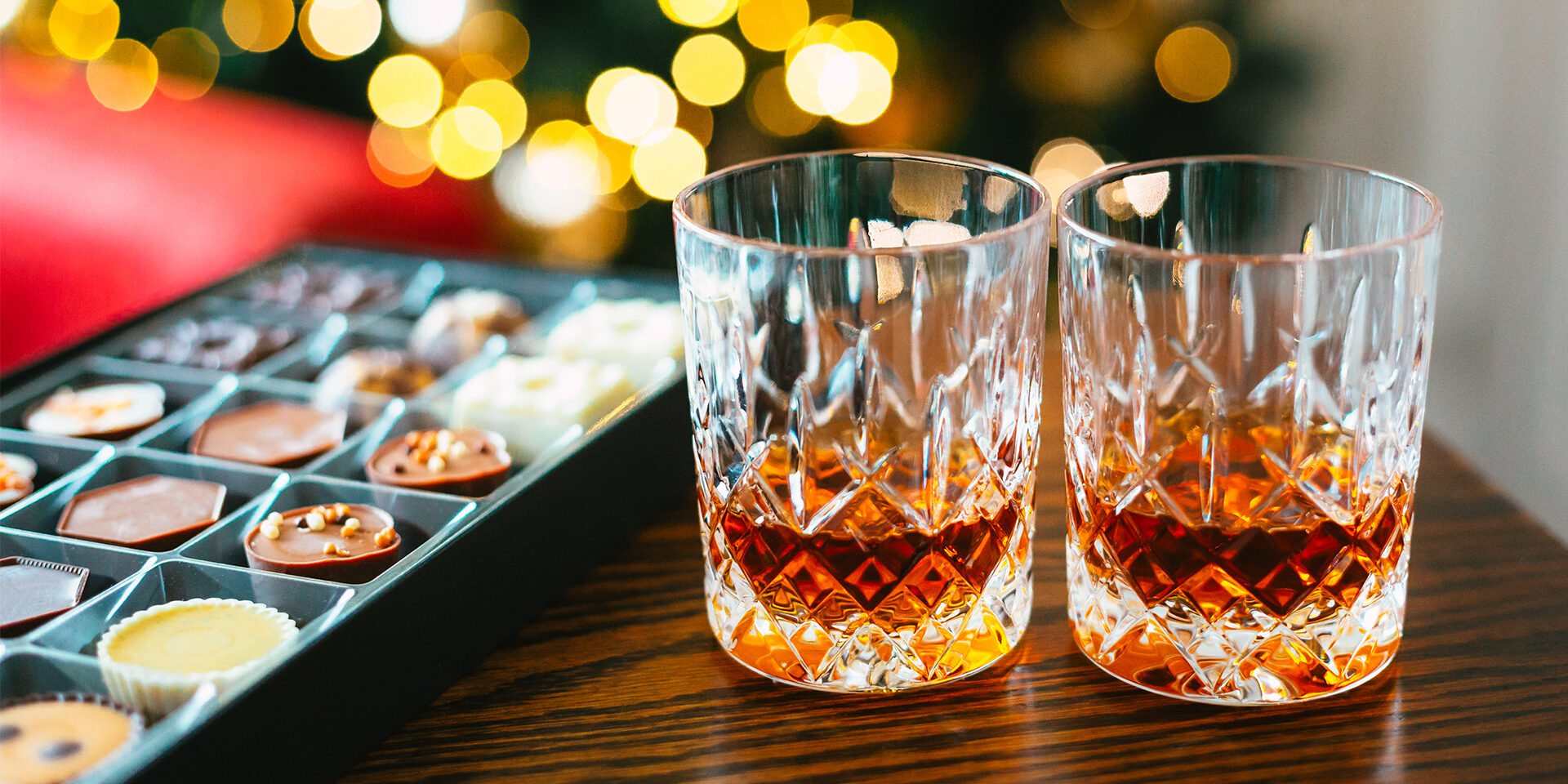 Close up color image depicting a box of gourmet chocolate truffles of different shapes and different varieties of chocolate. There are two crystal glasses filled with whisky. Selective focus with room for copy space.