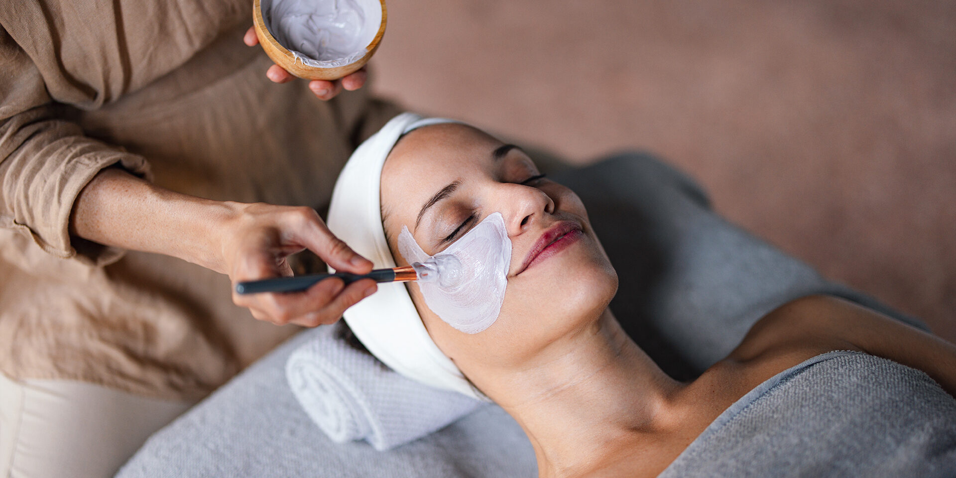 Beautiful Woman Enjoying Receiving a Facial Treatment at the Spa Close up shot of a beautiful smiling woman lying with her eyes closed while another woman is applying a face mask on her in the spa center. She is relaxed and enjoys the treatment.