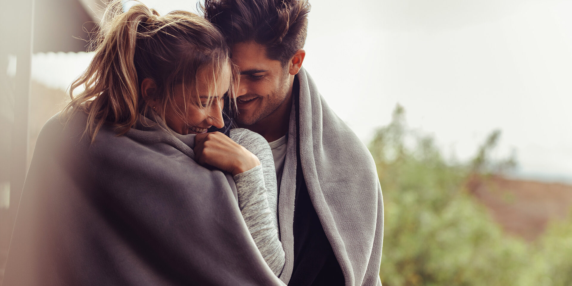 Romantic couple on a winter holiday. Man and woman standing together in a hotel room balcony wrapped in blanket. Couple embracing and smiling.