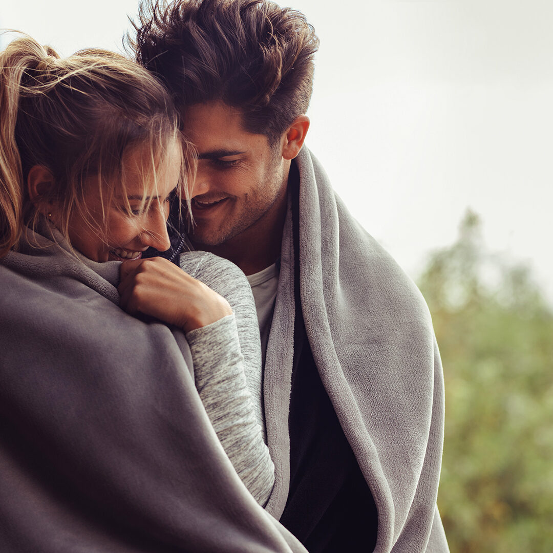 Romantic couple on a winter holiday. Man and woman standing together in a hotel room balcony wrapped in blanket. Couple embracing and smiling.