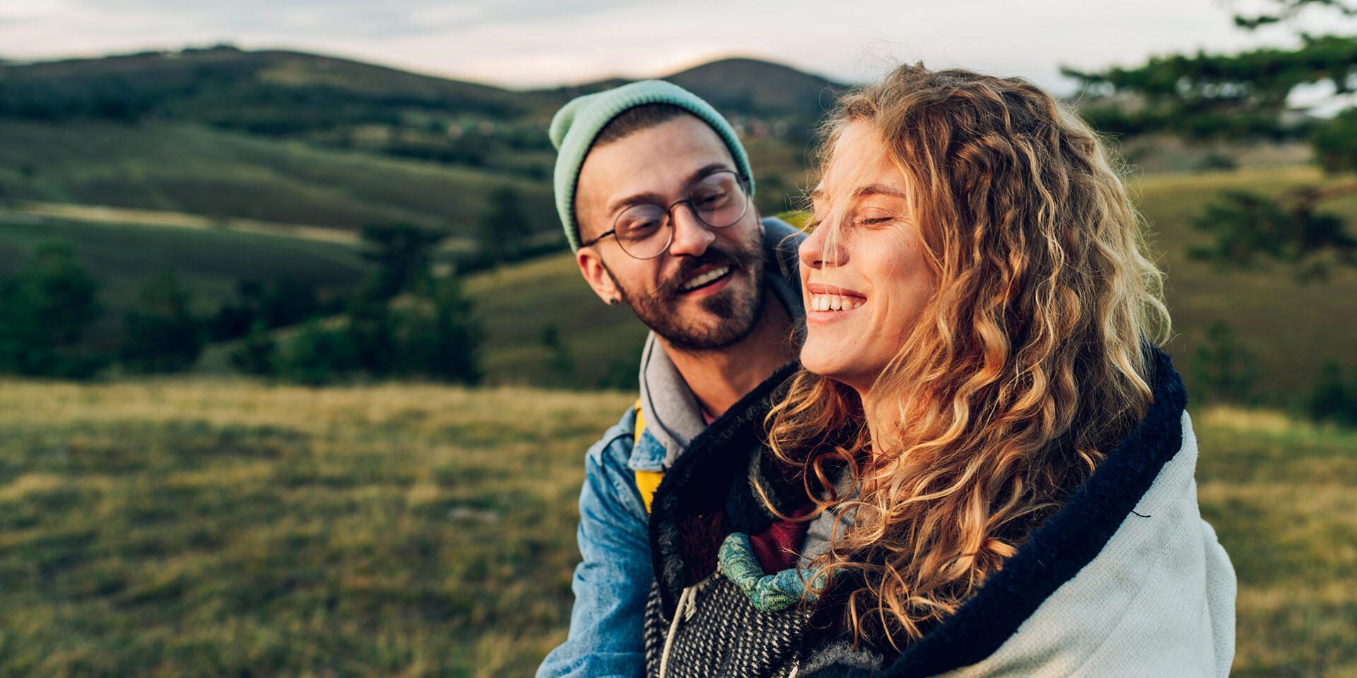 Couple of hikers hugging while walking on a mountain trail during a vacation Happy couple in love embracing and following trail along grassy mountain ridge and having fun together while walking in the nature. Healthy lifestyle and romantic getaway. Focus on a smiling woman..