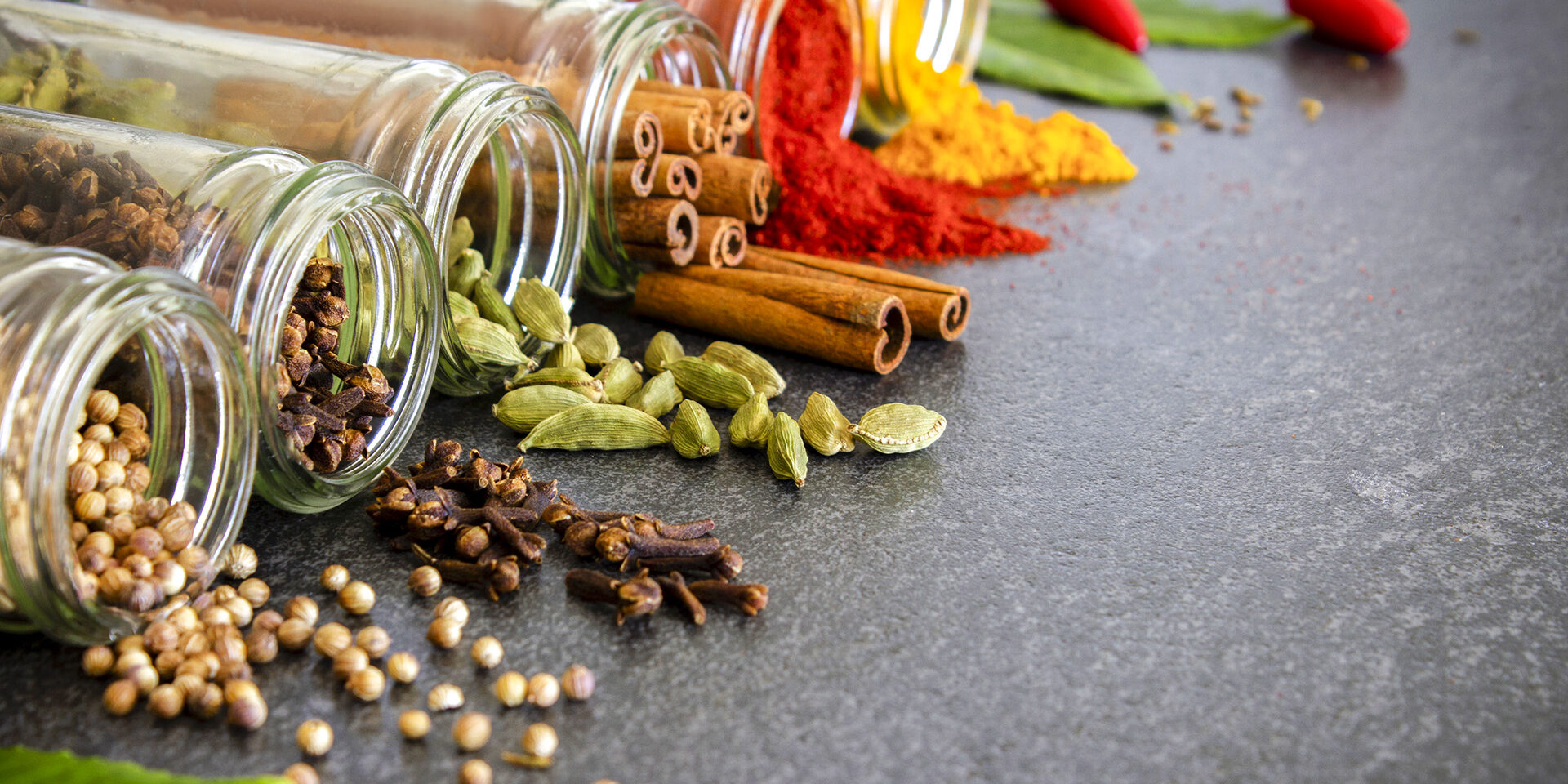 Jars of spices A selection of dried spices and herbs spilling out from glass jars onto a dark slate background.
Coriander seed, cloves, cardamom, cinnamon sticks, chilli powder, turmeric and fresh bay leaves.
Low angle view with copy space to the right of the image.
Close up.