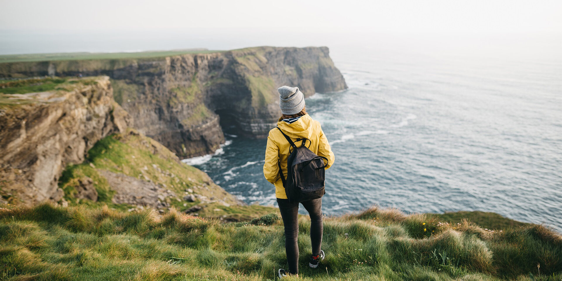 Young backpacker girl standing on the  Cliffs of Moher in yellow jacket with backpack