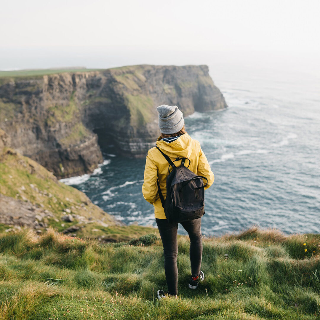 Young backpacker girl standing on the  Cliffs of Moher in yellow jacket with backpack