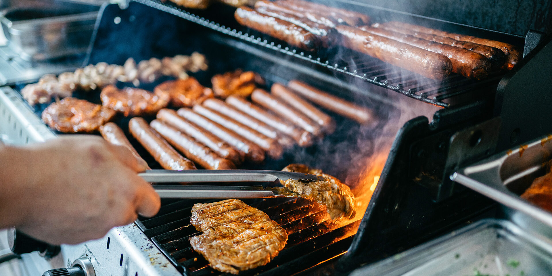 Grilling Meat and Hot Dogs on Gas Barbecue Grill Close-up view of various meats being grilled on a modern gas barbecue. The image shows juicy steaks and hot dogs cooking on hot grates with visible smoke and flames. A hand with tongs is visible, turning a perfectly seared steak. The composition captures the essence of outdoor cooking with multiple levels of the grill being utilized, showcasing both raw and cooking stages of the meat. Fresh herbs are visible in the background, adding to the culinary atmosphere.