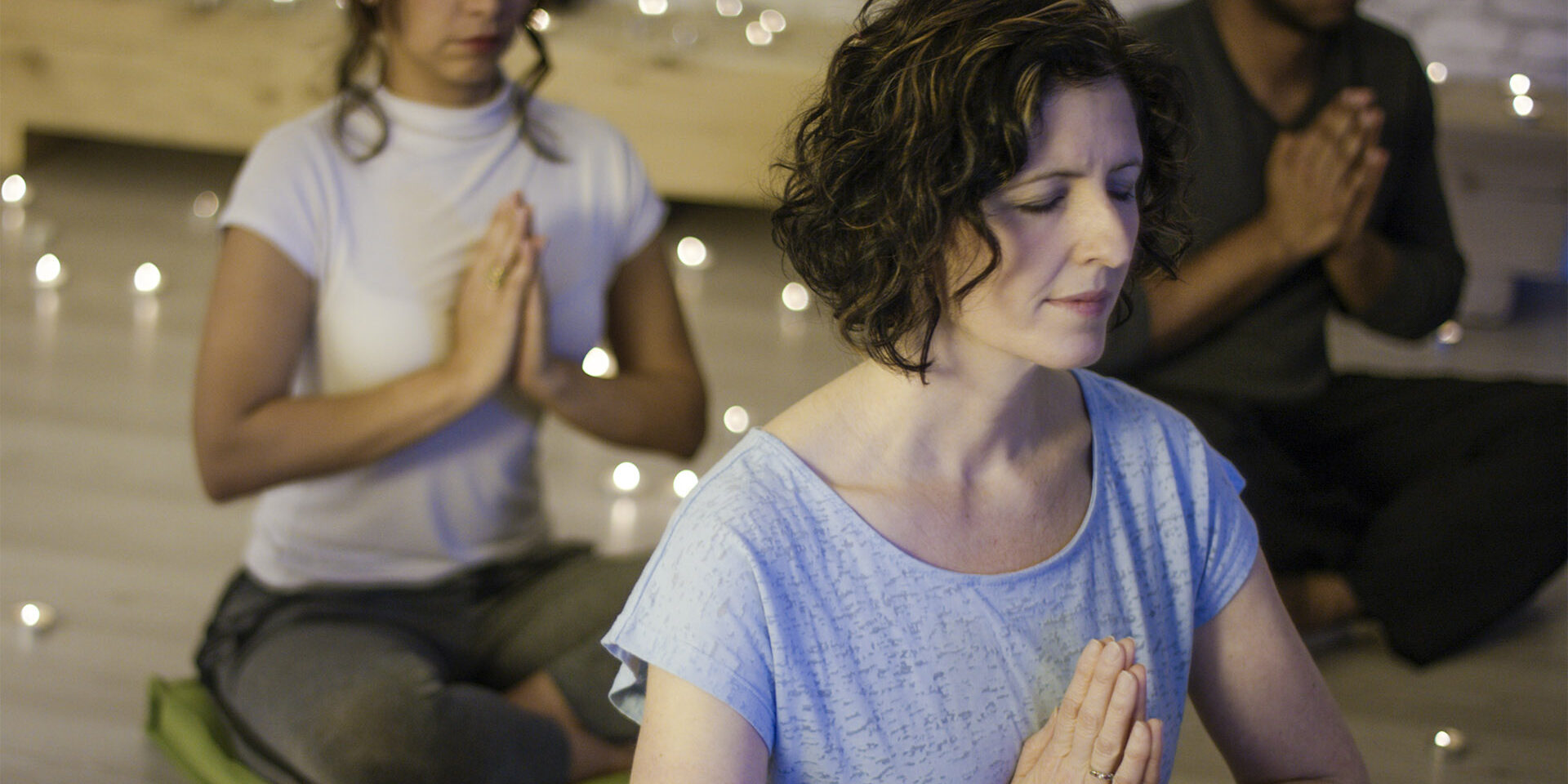 Mindfulness Beautiful young woman with short brown hair wearing a grey t-shirt meditating in prayer pose with a small multi-ethnic group of people during a meditation class surrounded by candles in a dimly lit room.