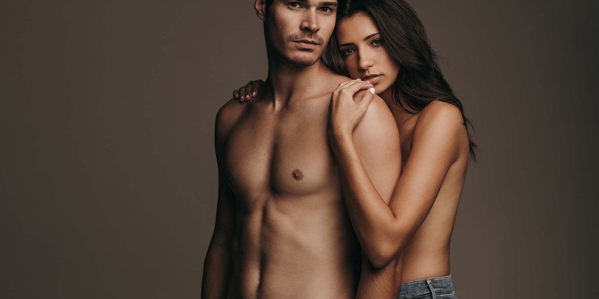 Beautiful young couple only wearing jeans standing together on brown background. Sensual man and woman looking at camera.