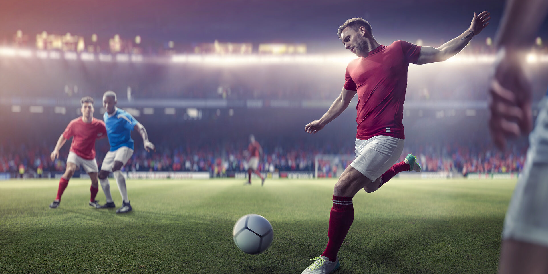 A professional male soccer player with foot back and arms out about to kick football in a volley during a soccer match. The player is dressed in generic red and white soccer kit and is near other players who are playing in a game on a generic outdoor soccer pitch.