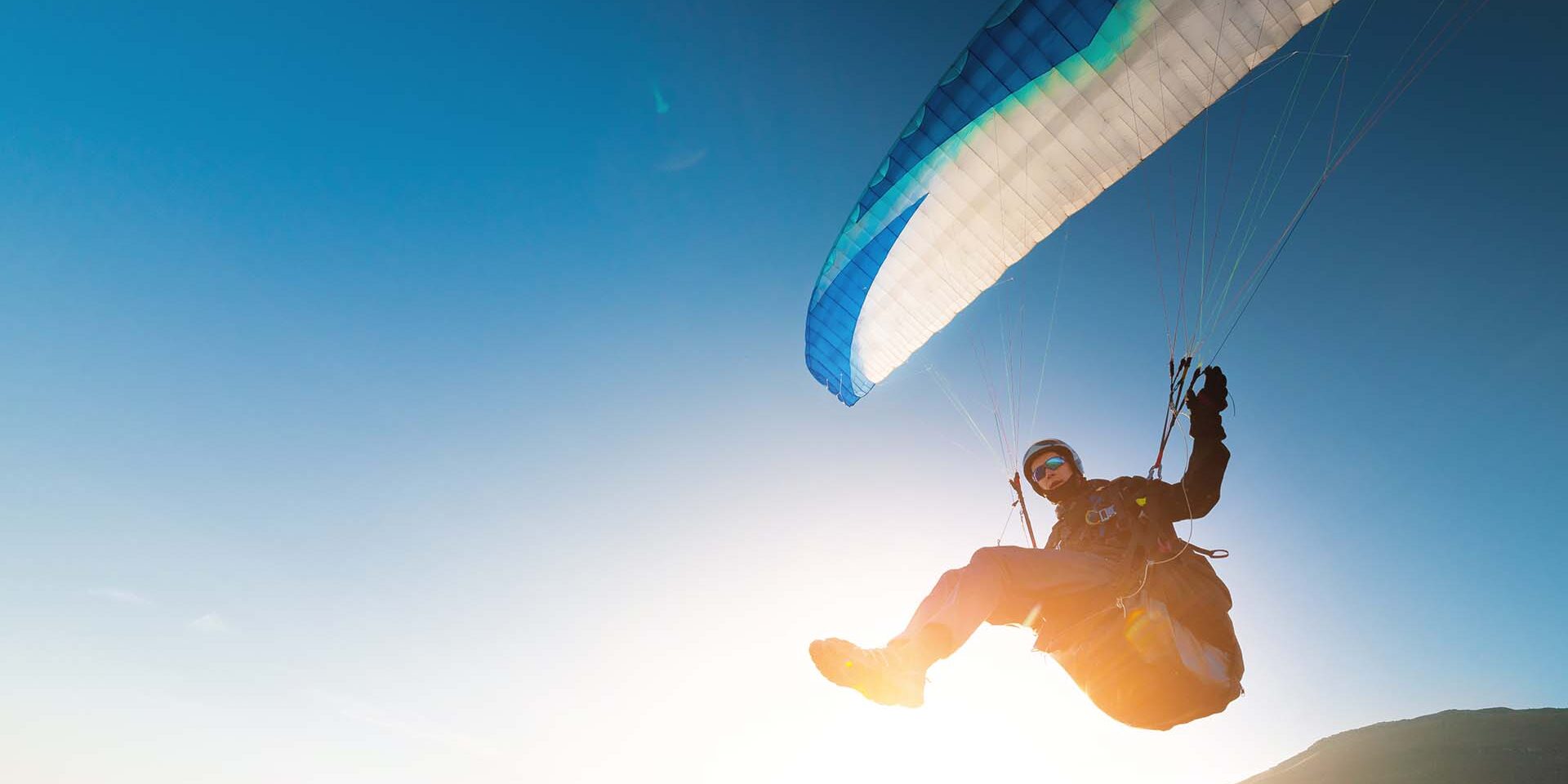 A paraglider takes off from a mountainside with a blue and white canopy and the sun behind. A paraglider is a silhouette. The glider is sharp, with little wing movement. A male paraglider launches a paraglider into the air.