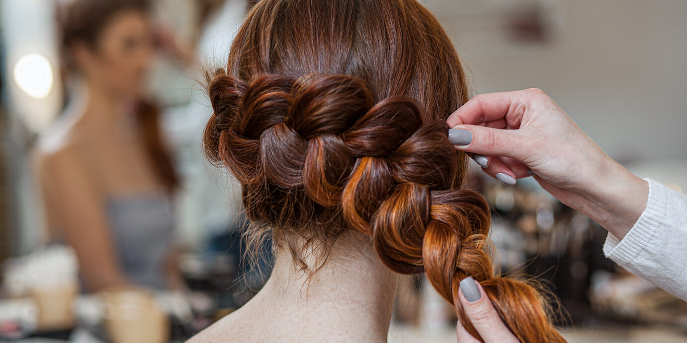 Beautiful, with long, red-haired hairy girl, hairdresser weaves a French braid, close-up in a beauty salon. Professional hair care and creating hairstyles.